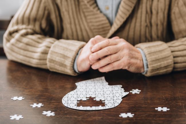 Starý člověk v hnědém svetru sedí za hnědým stolem u puzzle mozku, brain health